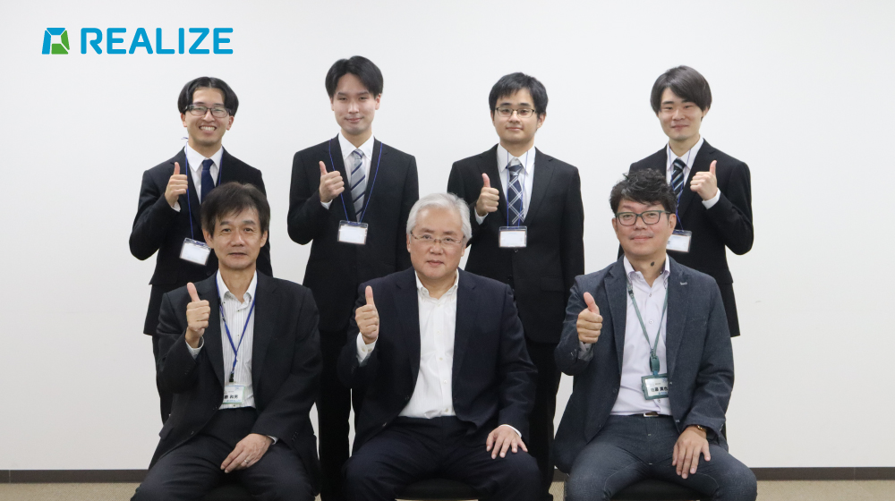 REALIZEの内定式後の記念撮影。役員と内定者が笑顔で親指を立てるポーズ。