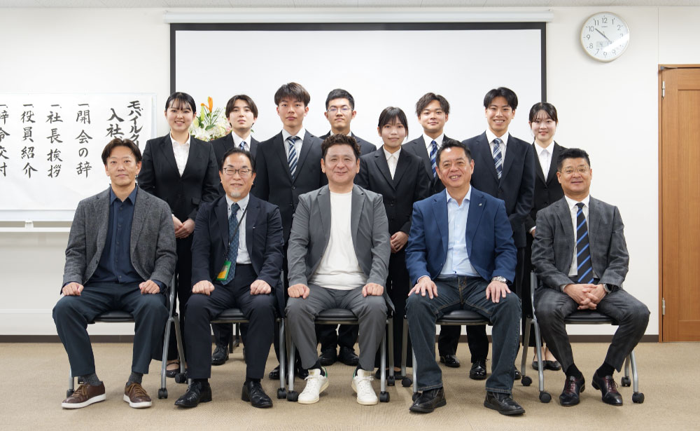 モバイルクリエイト株式会社の入社式で、役員と新入社員が並び記念撮影を行う様子。